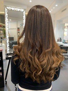 Una persona con lunghi capelli castani ondulati con riflessi più chiari, ripresa di spalle in un salone di bellezza.