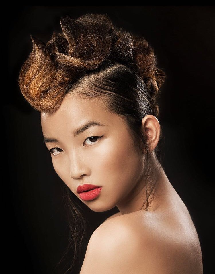 Ritratto di una persona con capelli scolpiti e strutturati, rossetto rosso acceso ed espressione neutra su uno sfondo scuro.
