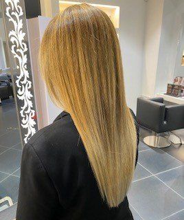 Una persona con lunghi capelli lisci color biondo miele, tagliati a V, vista di spalle in un salone da parrucchiere.