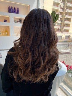 Capelli lunghi, mossi, castano scuro con riflessi più chiari e delicati, visti da dietro in un luminoso salone di bellezza al chiuso.
