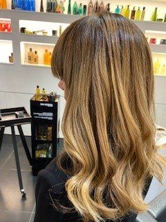 Una persona con capelli mossi, lunghi fino alle spalle, con balayage castano e biondo e frangia, vista di spalle in un salone da parrucchiere.