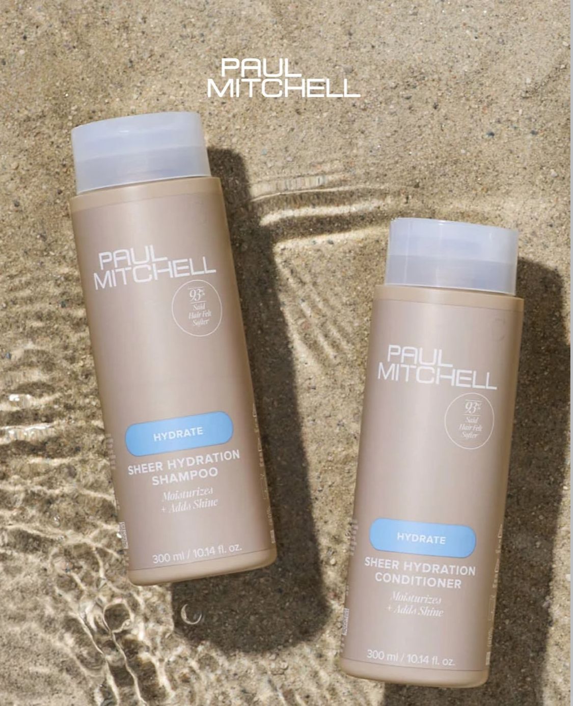 Due flaconi di shampoo e balsamo Paul Mitchell Super Hydration poggiano su un terreno sabbioso parzialmente coperto da acqua bassa.