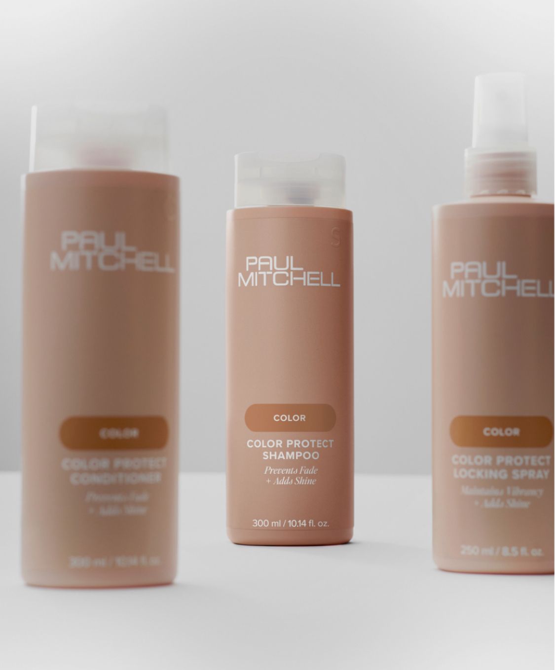Tre flaconi color cuoio di prodotti per la cura dei capelli Paul Mitchell, contenenti shampoo, balsamo e spray fissante, appoggiati su una superficie bianca.