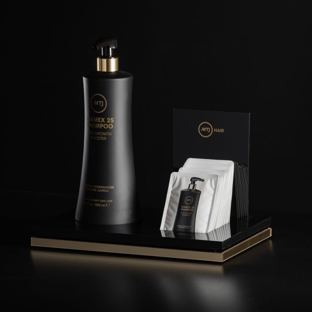 Una bottiglia di shampoo nera è posizionata accanto a un piccolo espositore di campioni di prodotto bianchi su una base a più livelli nera e dorata.
