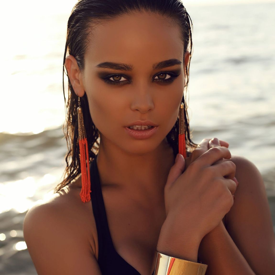 Una donna con i capelli bagnati indossa vistosi orecchini a nappe arancioni e un bracciale rigido dorato, in posa davanti all'oceano al tramonto.