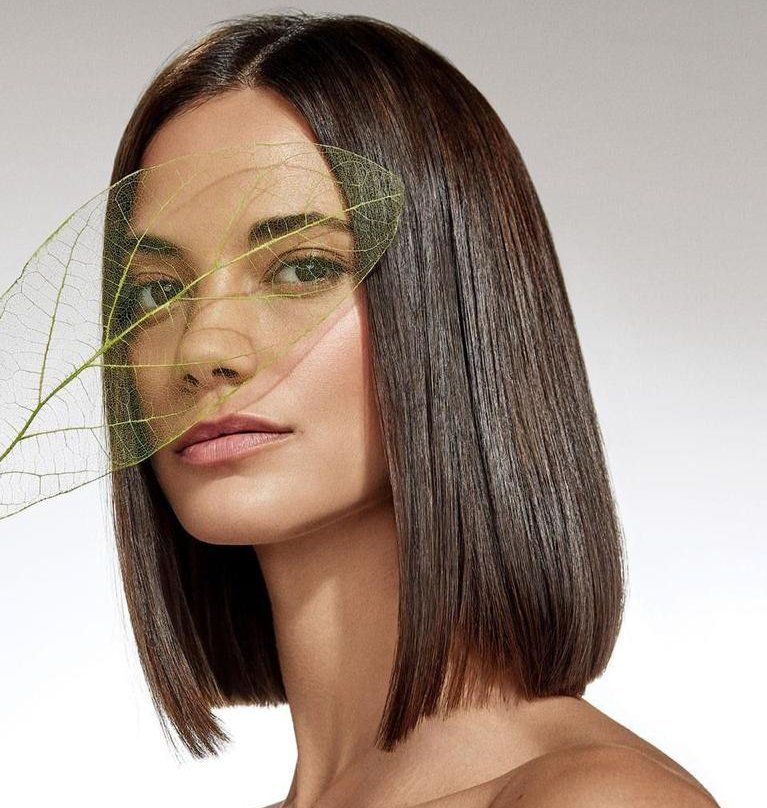 Una persona con i capelli castani lunghi fino alle spalle posa tenendo una foglia verde traslucida su un lato del viso.