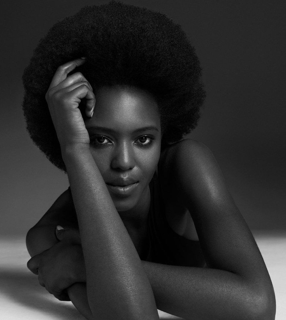 Una persona con una chioma afro naturale appoggia il mento sulla mano, guardando direttamente in camera in una luce soffusa.