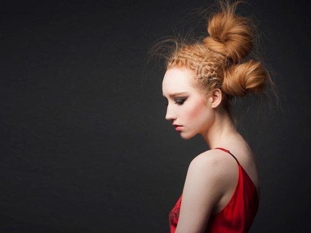 Ritratto di profilo di una persona con capelli rossi intrecciati in modo elaborato e raccolti in due chignon sulla sommità del capo, che indossa una maglietta rossa su sfondo nero.
