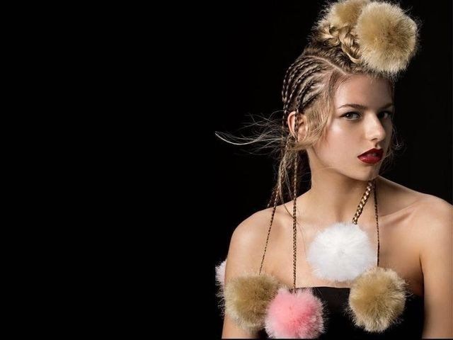 Una persona con i capelli intrecciati e grandi pon-pon colorati di pelliccia sintetica attaccati ai capelli e al collo, su sfondo nero.
