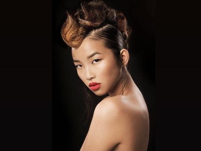 Una persona con un'acconciatura elaborata e d'effetto, rossetto rosso e spalle scoperte posa davanti a uno sfondo nero uniforme.