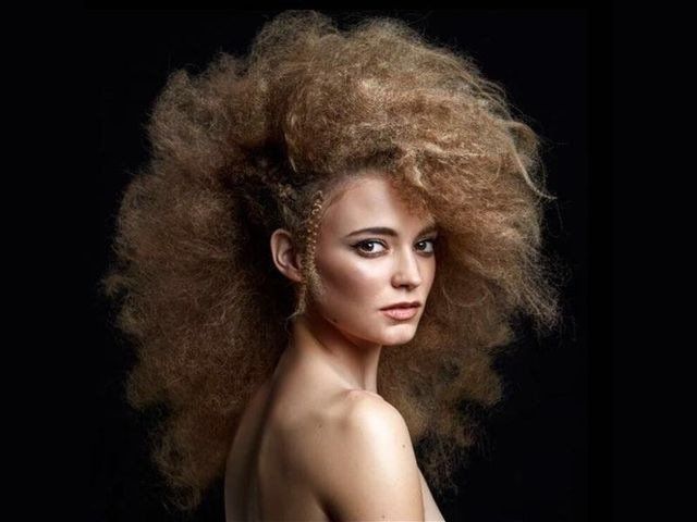 Una persona con capelli castano chiaro, voluminosi e strutturati, acconciati verso l'alto, che guarda indietro oltre la spalla, su uno sfondo nero.