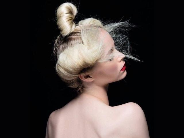 Una persona con i capelli biondo platino raccolti in uno chignon alto e voluminose ciocche laterali posa davanti a uno sfondo nero uniforme.