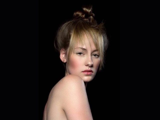 Una persona con i capelli chiari raccolti in uno chignon e la frangia posa davanti a uno sfondo nero, guardando verso la macchina fotografica.