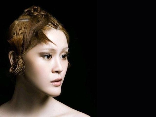 Una persona con i capelli acconciati in trecce elaborate e un trucco leggero, su uno sfondo nero uniforme, guarda verso sinistra.