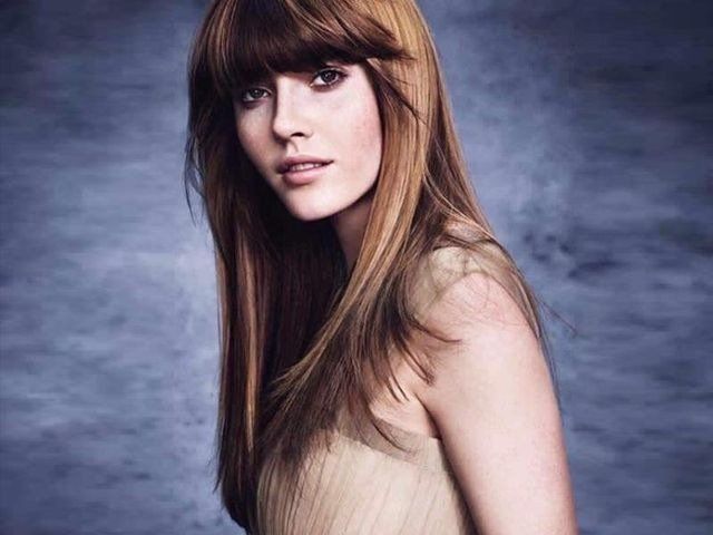 Una persona con lunghi capelli castani scalati e frangia, che indossa una maglietta color cachi su uno sfondo blu sfocato e texturizzato.