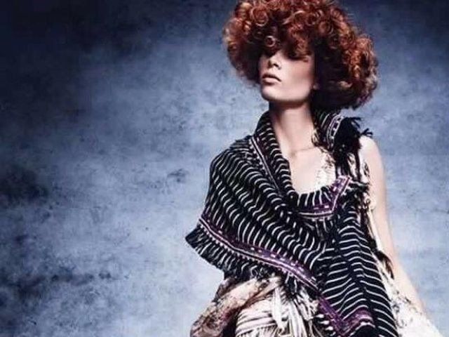 Una persona con capelli ricci castano-rossicci indossa uno scialle scuro a righe con frange su uno sfondo grigio-blu dalla trama particolare.