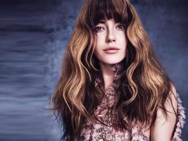 Una persona con lunghi capelli castani ondulati con mèches e frangia posa davanti a uno sfondo blu scuro con texture.