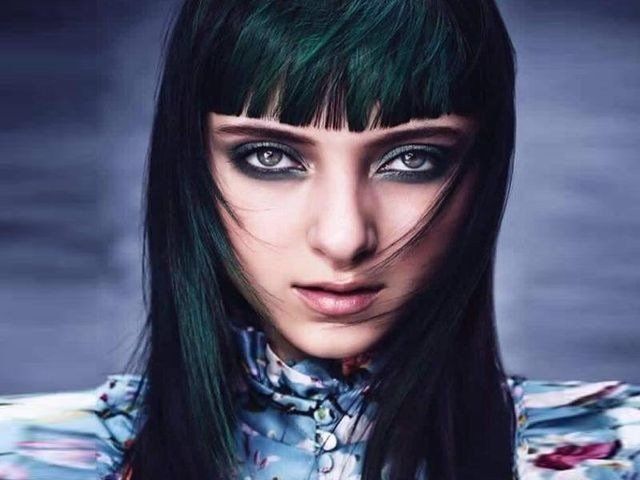 Ritratto di una persona con capelli tinti di verde scuro e frangia dritta, che indossa un trucco occhi vistoso e un colletto a fantasia floreale.