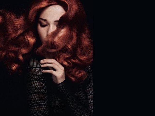Una persona con una folta chioma ondulata color rame che le copre il viso, indossa un top scuro e strutturato su uno sfondo nero.
