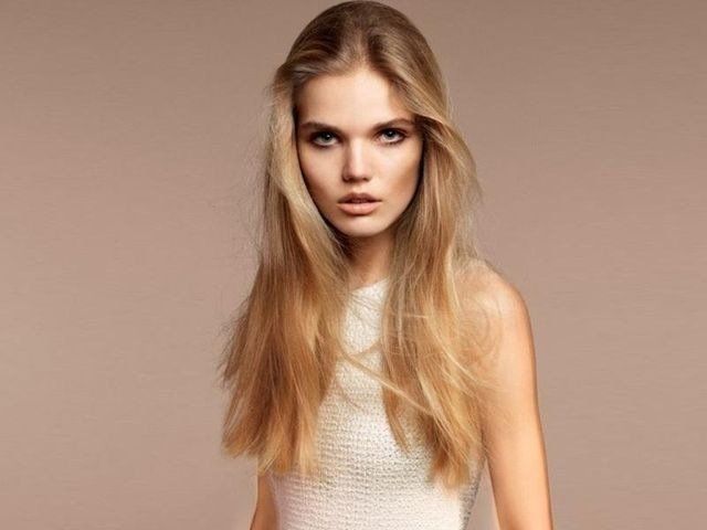 Una persona con lunghi capelli biondi e ondulati guarda direttamente in camera, su uno sfondo neutro color beige.