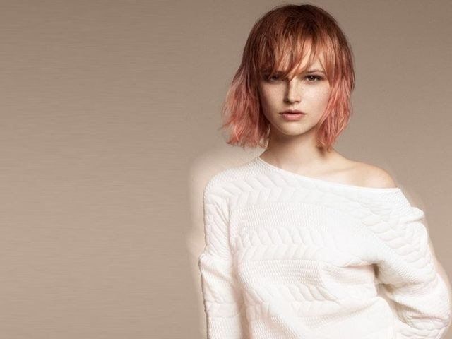 Una persona con capelli mossi color pesca e frangia indossa un maglione di maglia bianco a spalle scoperte su uno sfondo beige.