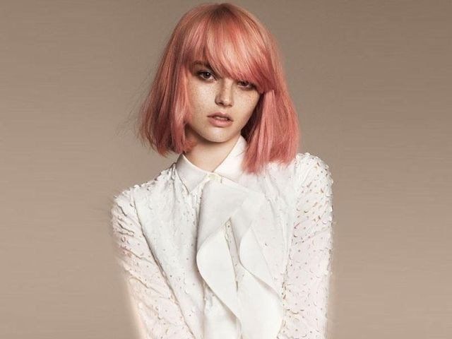 Una persona con capelli rosa pastello lunghi fino alle spalle e frangia, che indossa una camicetta bianca di pizzo con volant su uno sfondo semplice.