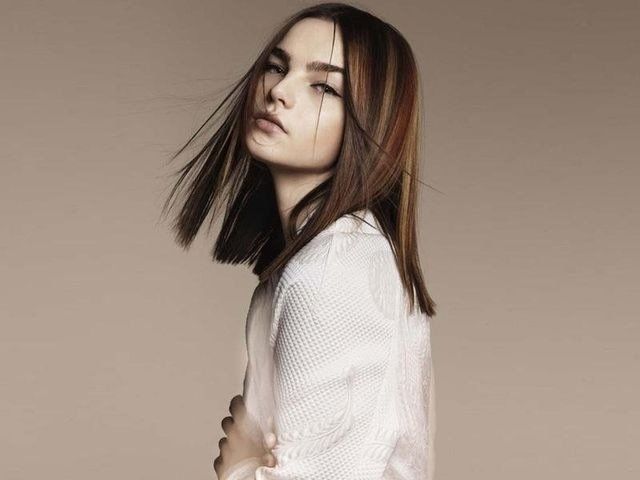 Una persona con i capelli castani di media lunghezza posa indossando una maglietta bianca testurizzata su uno sfondo beige uniforme.