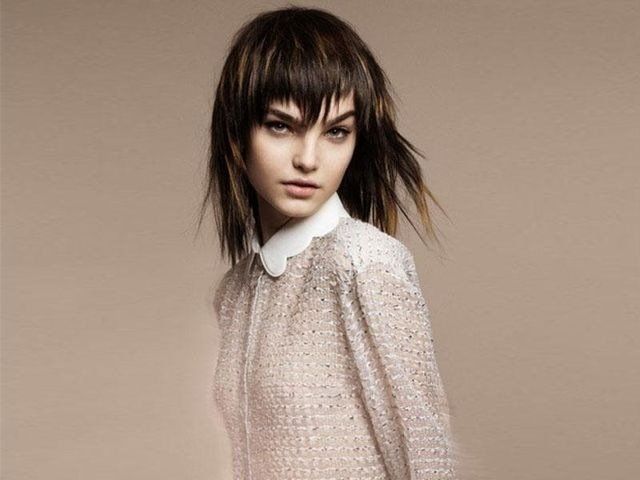 Una persona con un taglio di capelli scuro, scalato e con la frangia, che indossa una camicetta bianca con colletto, posa su uno sfondo color beige.