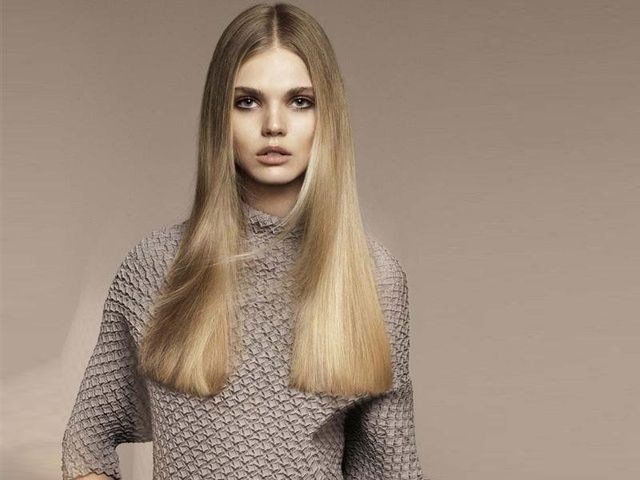 Una persona con lunghi capelli biondi e lisci che indossa un maglione grigio a collo alto con trama, su uno sfondo semplice e dai toni chiari.