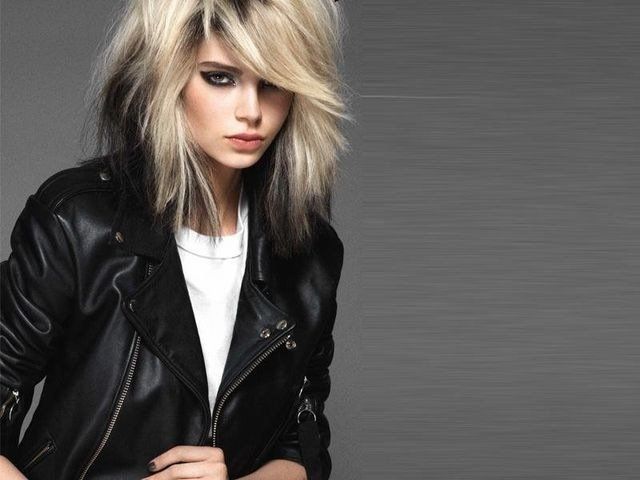 Persona con capelli biondi e scuri scalati e trucco occhi audace, che indossa una giacca di pelle nera su sfondo grigio.