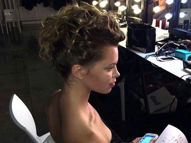 Una persona seduta su una sedia da trucco, con i capelli ricci, guarda un telefono in un camerino ben illuminato.