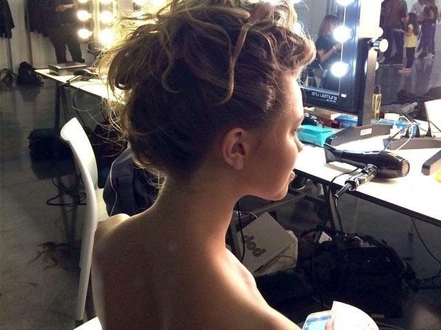 Una persona con i capelli ricci raccolti in uno chignon disordinato, vista di spalle in un camerino ben illuminato dietro le quinte.