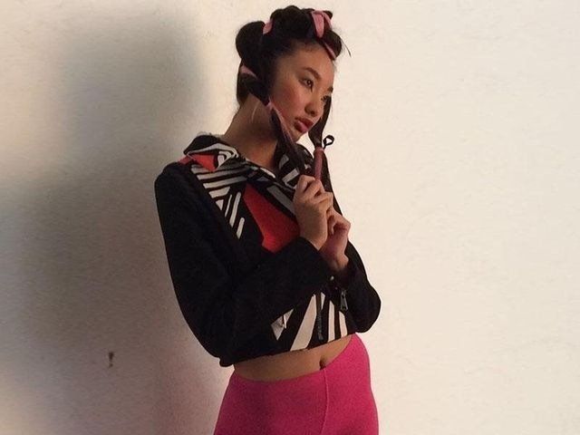 Una persona con i capelli raccolti in due codini, che indossa una giacca nera a fantasia e pantaloni rosa, posa davanti a uno sfondo bianco uniforme.