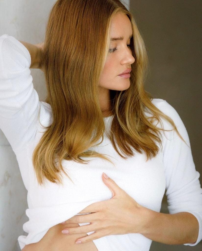 Una persona con i capelli biondi ondulati indossa una camicia bianca a maniche lunghe e tiene una mano sul ventre.