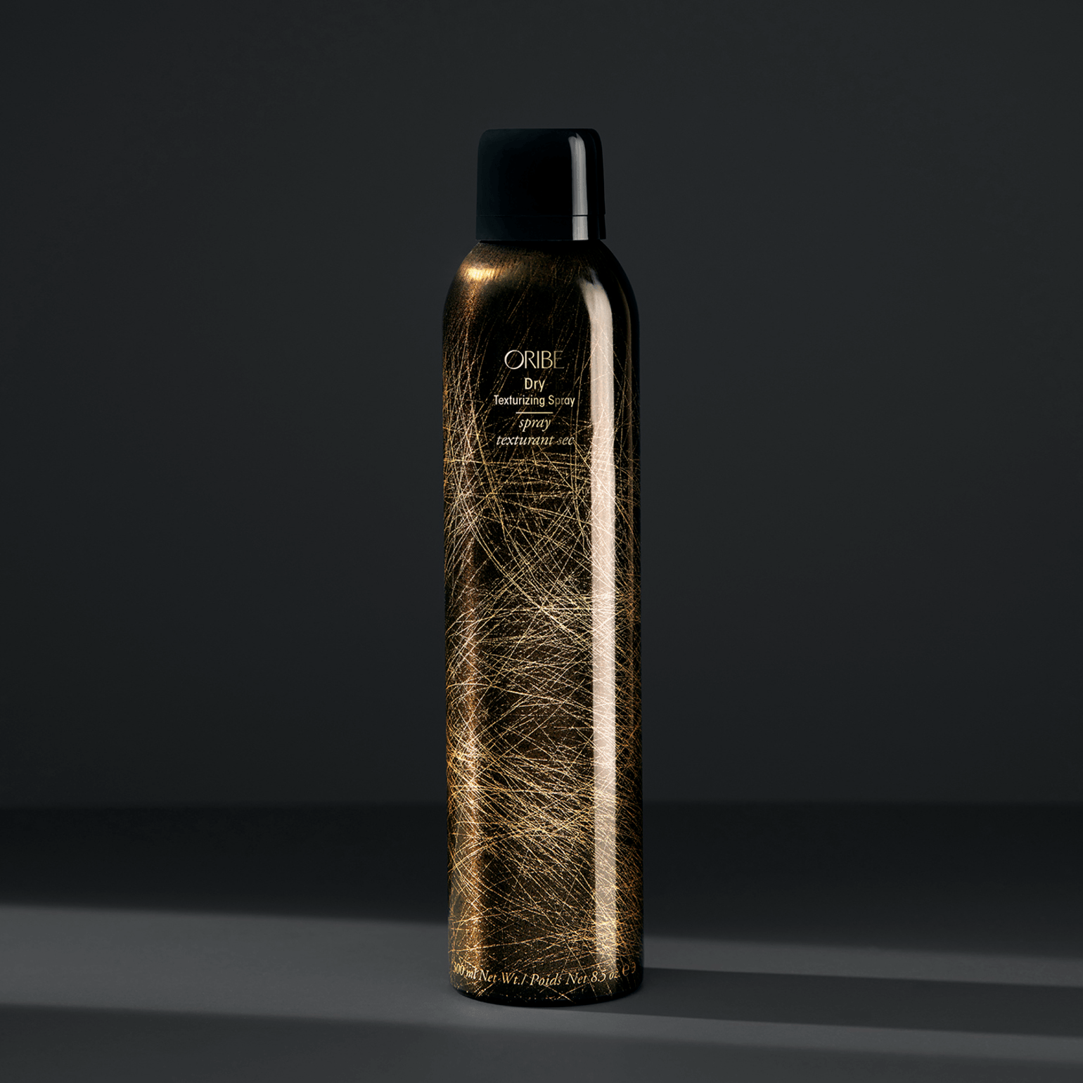 Un flacone spray texturizzante a secco Oribe con finitura dorata e tappo nero si staglia su uno sfondo grigio scuro uniforme.