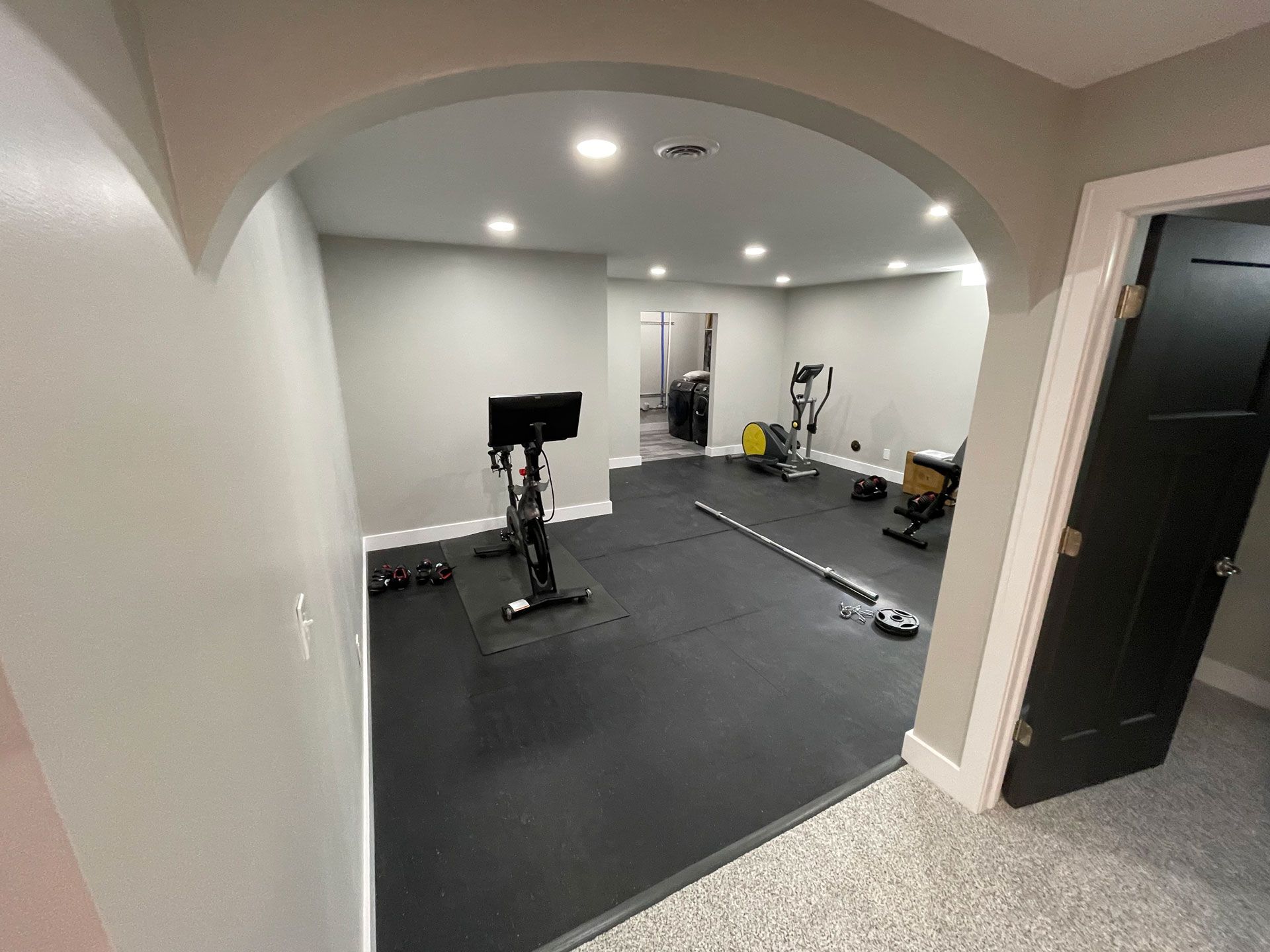 A home gym with black rubber flooring, an exercise bike, a cross-trainer, and weights, viewed through an arched doorway.