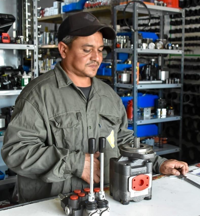 Una persona con uniforme gris y gorra trabajando en un banco de trabajo con componentes hidráulicos en un almacén de repuestos.