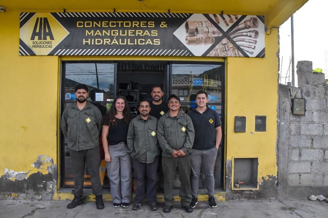 Seis personas posan frente a una tienda amarilla con un cartel que dice "CONECTORES & MANGUERAS HIDRAULICAS" encima de la entrada.