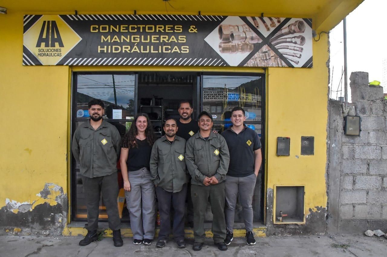 Seis personas posan frente a una tienda amarilla con un cartel que dice "CONECTORES & MANGUERAS HIDRAULICAS" encima de la entrada.