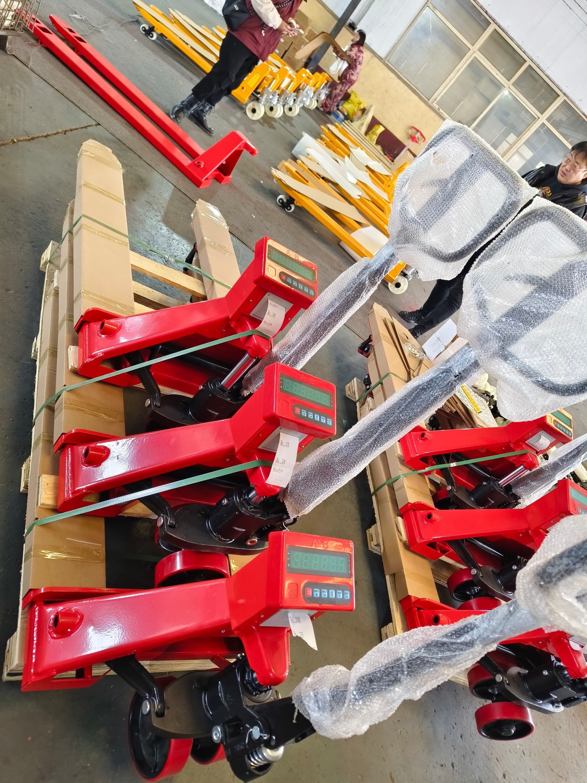 Varias transpaletas rojas con básculas digitales incorporadas, envueltas en plástico protector, se encuentran sobre palés de madera en un almacén.