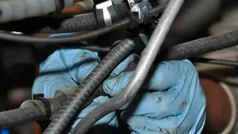 Unas manos con guantes protectores azules trabajan en un complejo arreglo de mangueras y cables de automóviles dentro del compartimiento del motor.