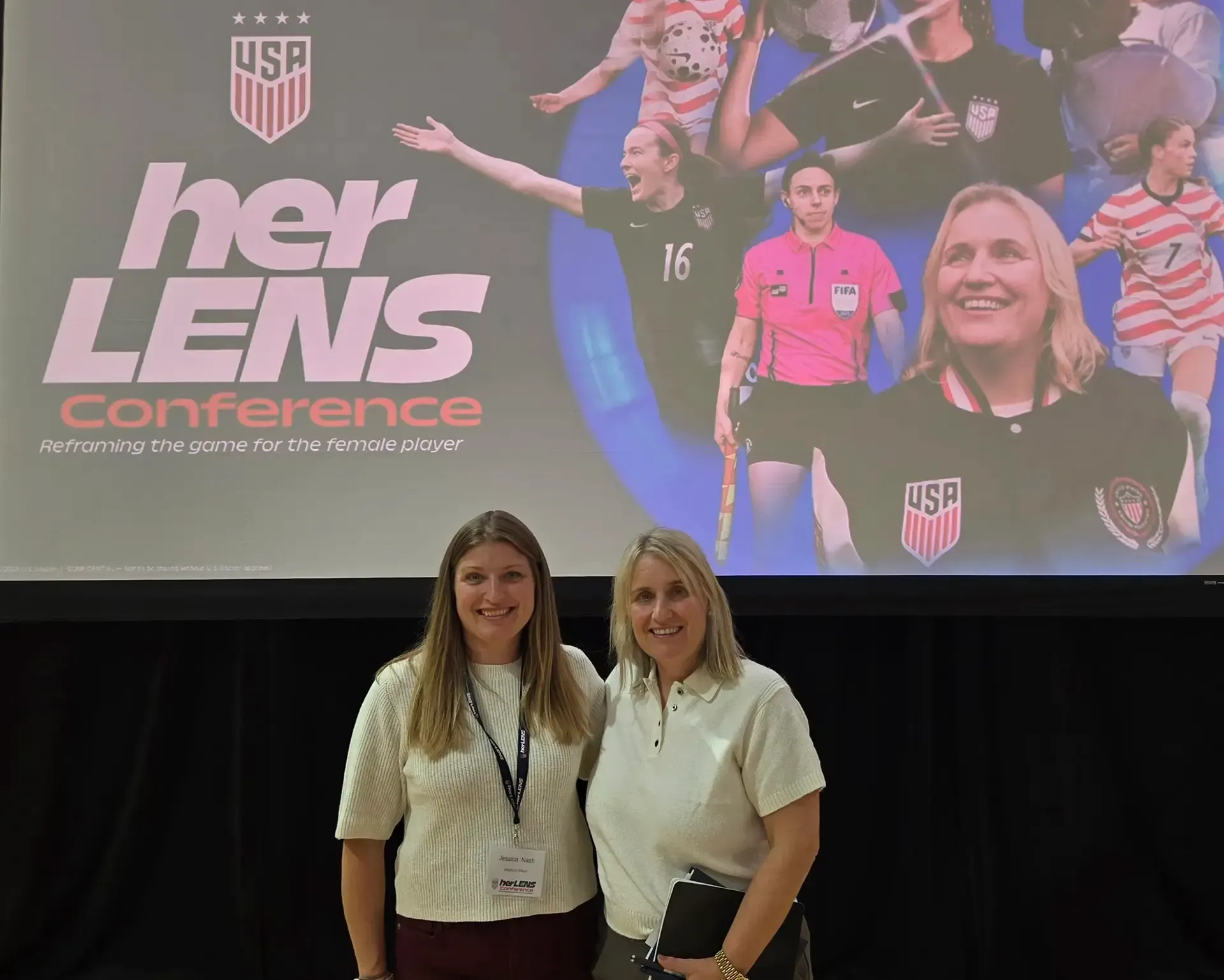 Two women standing in front of a “Her Lens Conference” stage backdrop with soccer imagery and USA logos