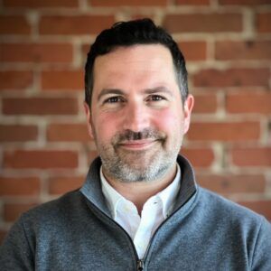 Smiling man in a gray zip sweater and white shirt against a brick wall background