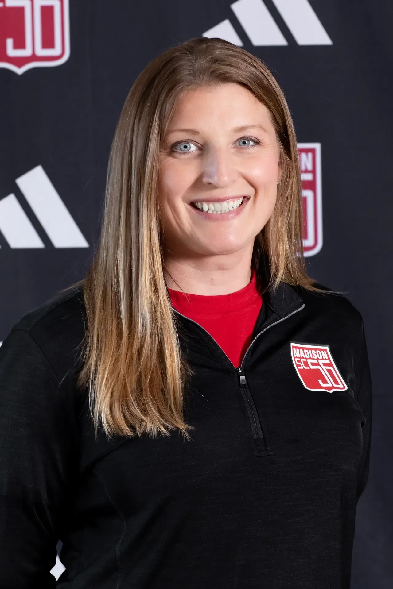 Smiling person in black zip-up jacket with red logo, standing before a branded backdrop