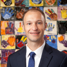 Portrait of a smiling man in a dark suit and blue tie in front of a colorful handprint mural