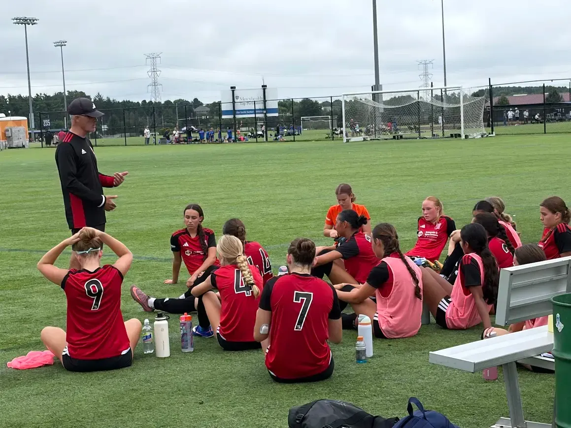 Youth soccer team in red jerseys listening to a coach on a field near a goal.