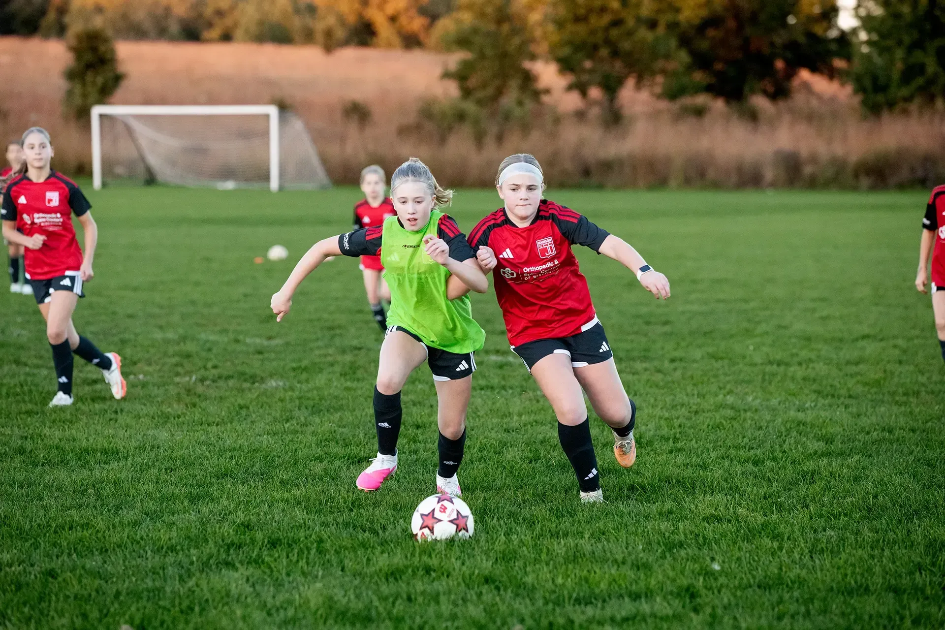 Two soccer players in red and green chase the ball on a grassy field, with a goal in the background