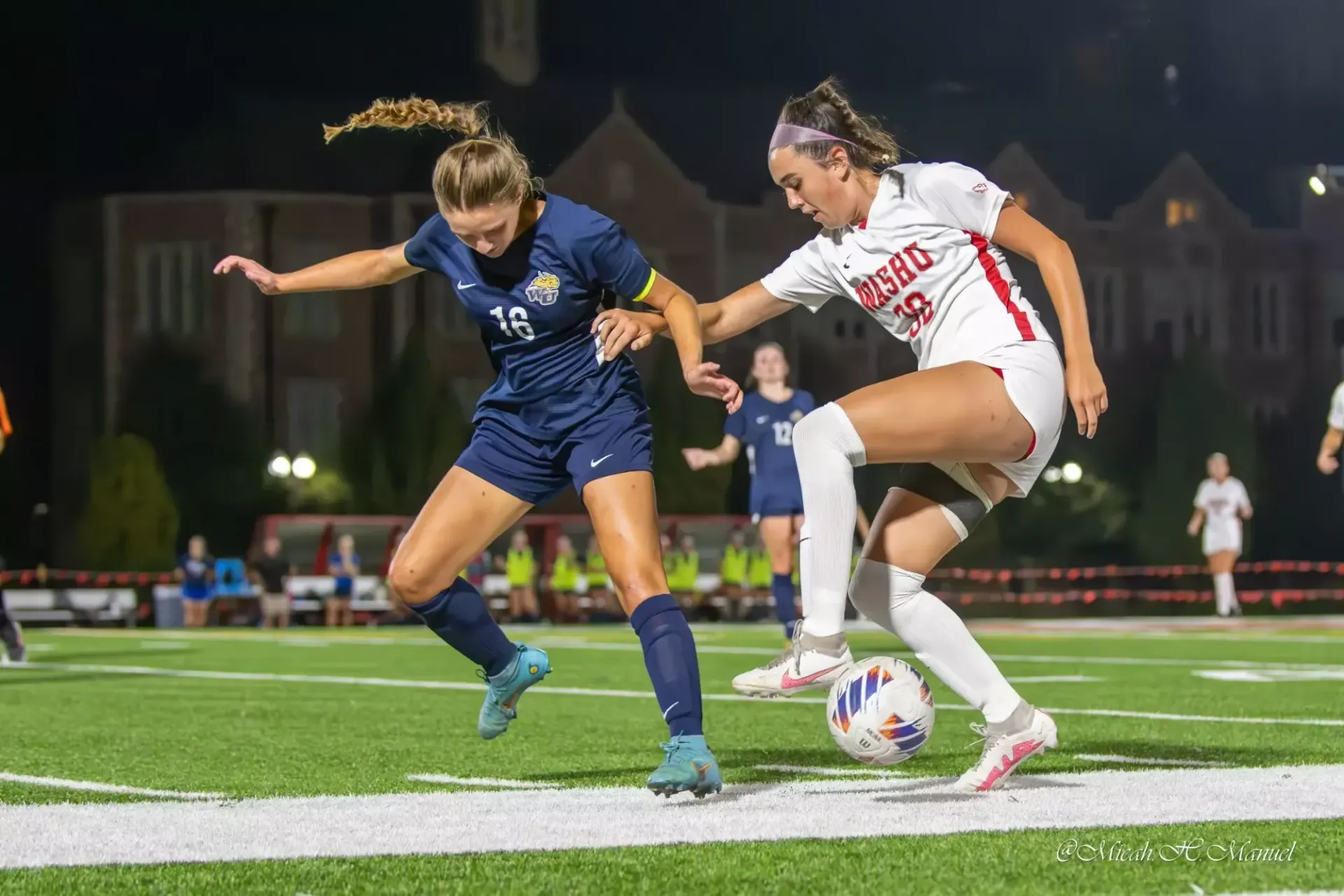 Two soccer players battle for the ball on a lit field at night, one in blue and one in white.