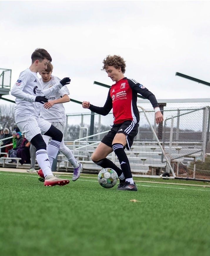 Three soccer players contest the ball on a green field, with a red-and-black player in front of two white-clad opponents.