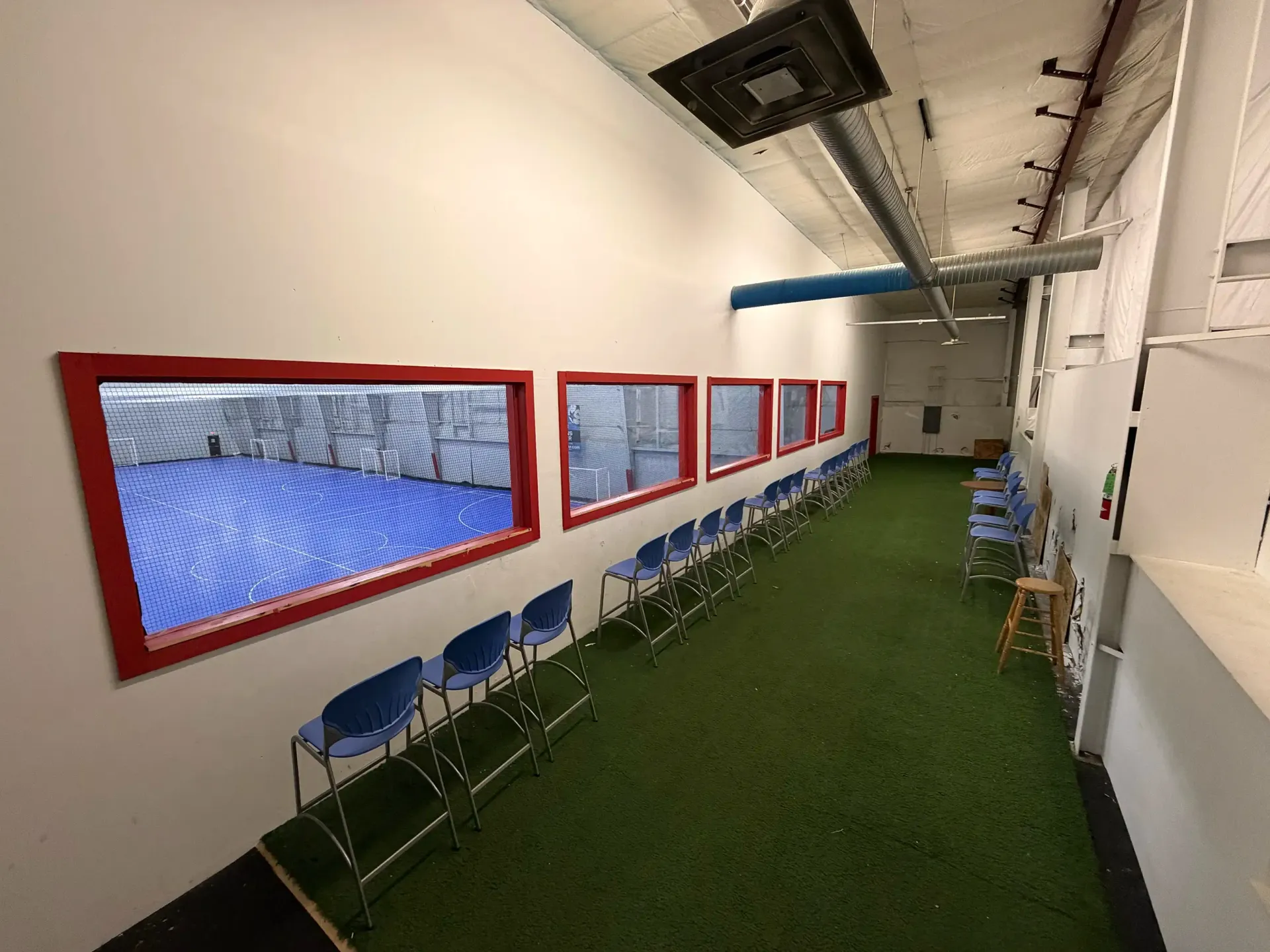 Narrow indoor hallway with green carpet, blue chairs, and red-framed window openings along the left wall